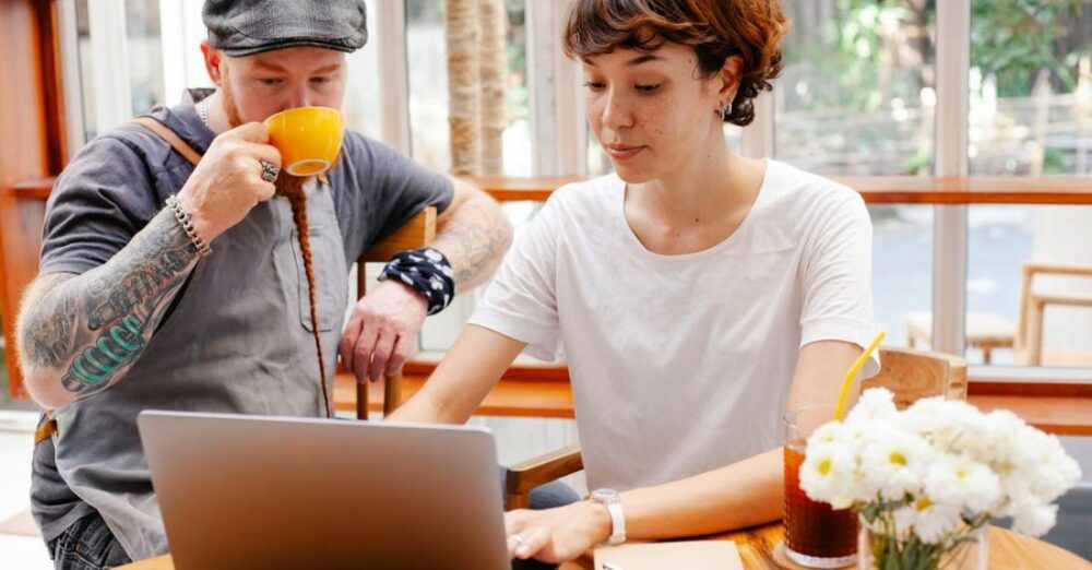 Remote Teams - Pensive young woman typing on netbook while man with cup drinking coffee at table of cafe